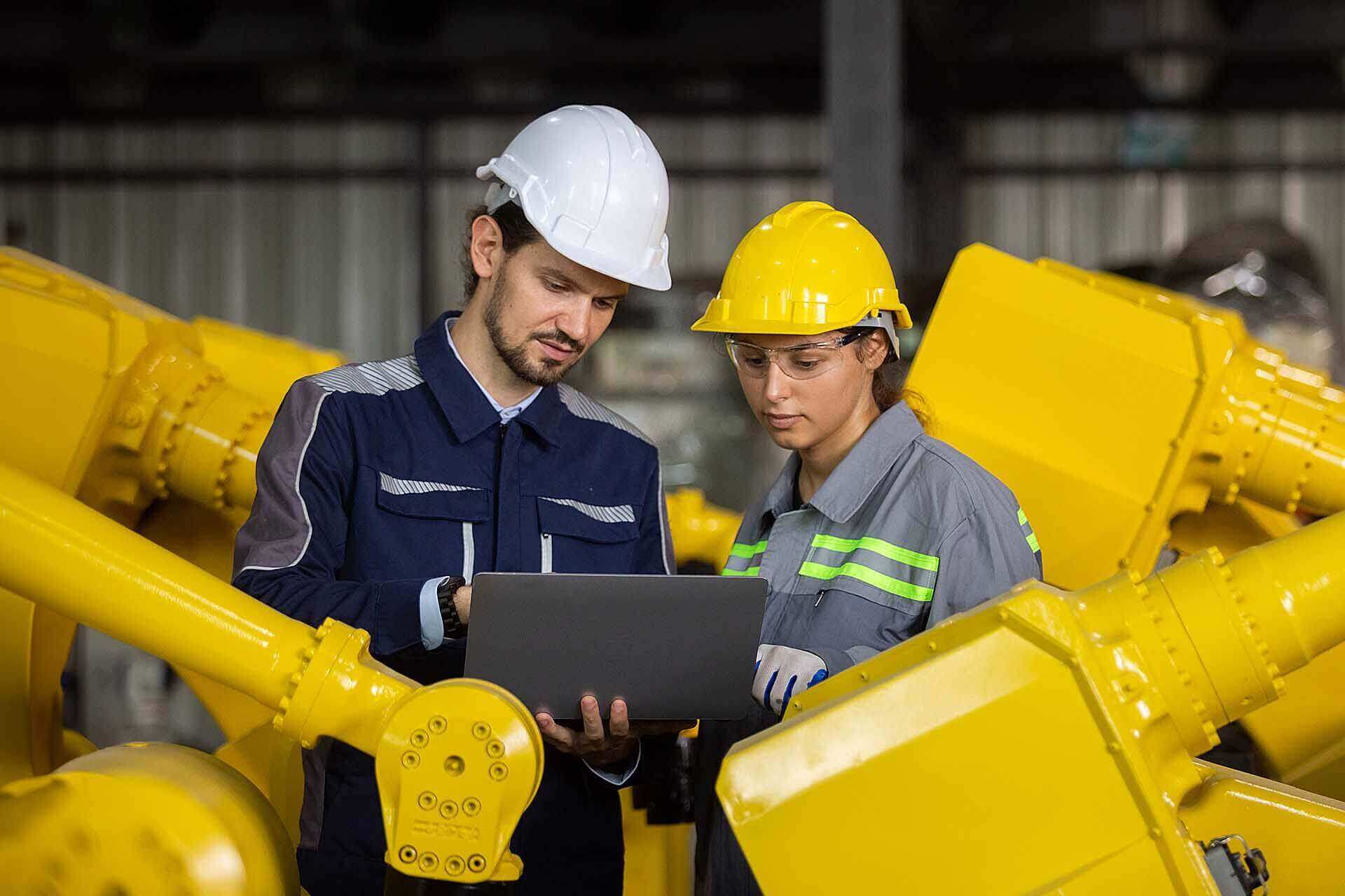 Two industrial workers reviewing information on a laptop in a factory with yellow machinery