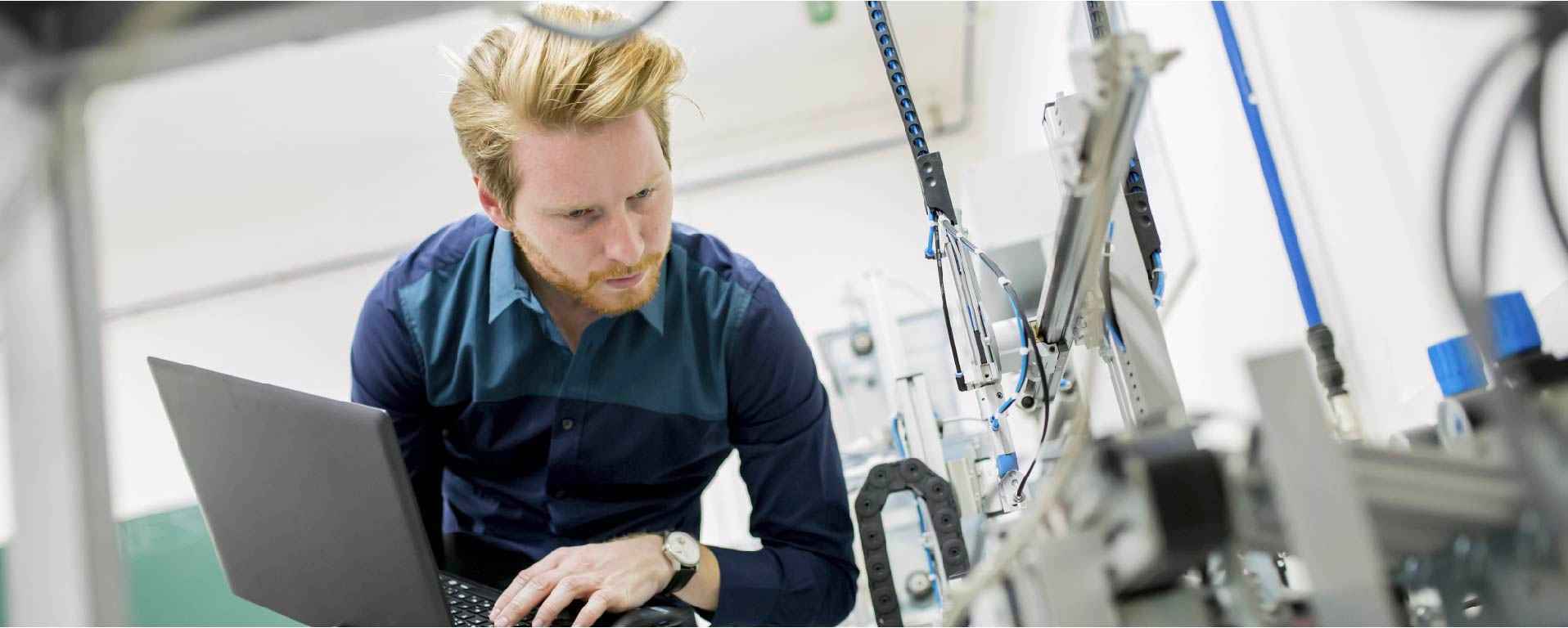 An engineer working on a laptop, closely inspecting machinery with connected cables and components in a lab setting