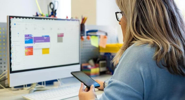 A person holding a smartphone looks at a computer screen displaying a digital calendar