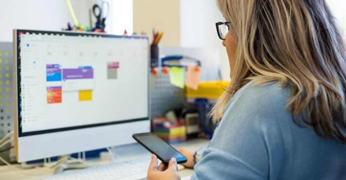 A person holding a smartphone looks at a computer screen displaying a digital calendar