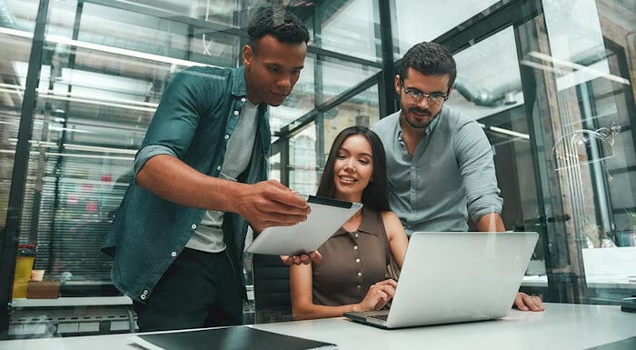 Three colleagues collaborating in a modern office, reviewing information on a tablet and laptop