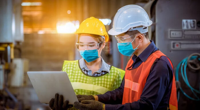 Two factory workers wearing safety helmets, face masks, and vests reviewing information on a laptop