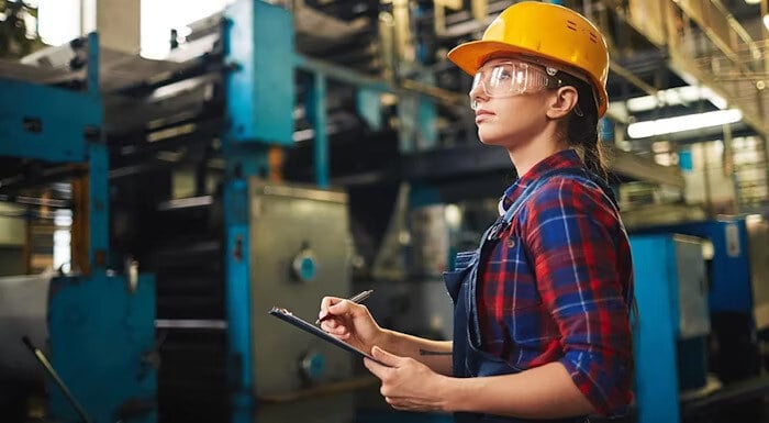 A female factory worker in a yellow hard hat and safety goggles holding a clipboard and inspecting machinery