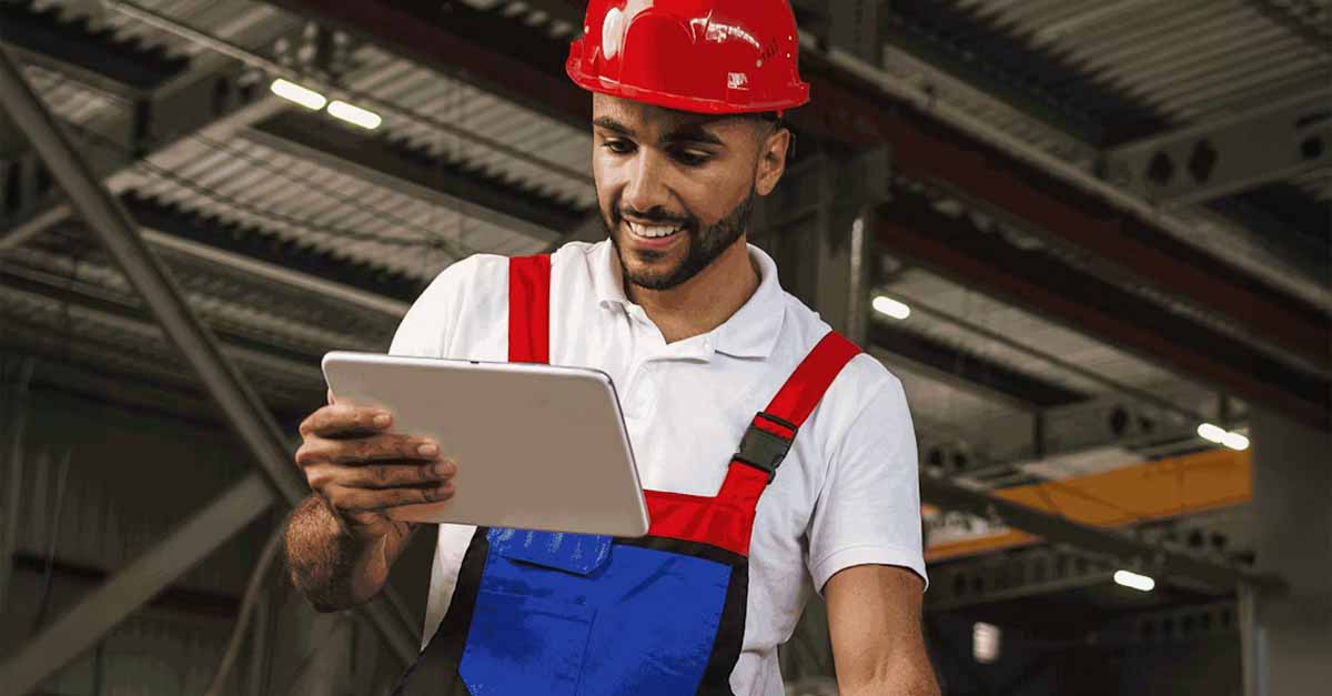 A smiling industrial worker in a red hard hat and overalls using a tablet in a factory setting