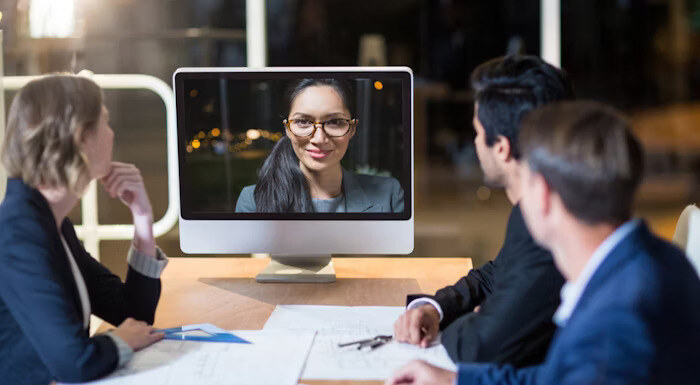 Three people in a meeting are sitting around a table, watching a woman on a computer screen during a video conference