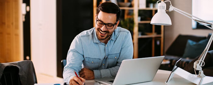 A smiling man sits at a desk with an open laptop, writing in a notebook while working in a well-lit office