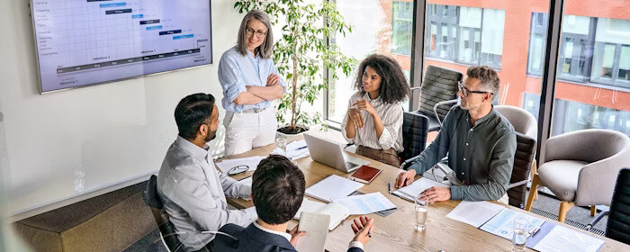 A group of colleagues sits around a conference table discussing a presentation displayed on a large screen in a modern office with windows