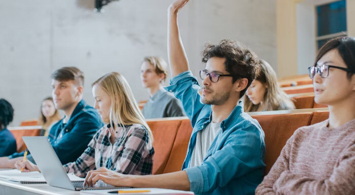 A student sitting in a lecture hall raises his hand while others around him take notes and use laptops