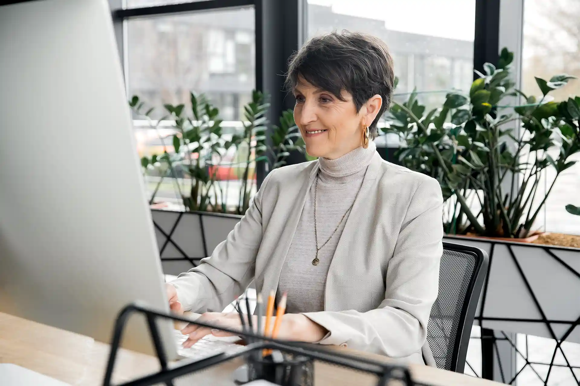 A woman in a light-colored blazer works at a computer in a modern office, with plants and large windows in the background