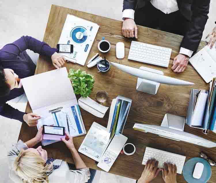 A top-down view of a group of people working at a desk, using computers, tablets, and reviewing documents with charts and graphs