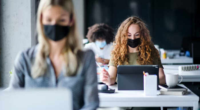 People wearing face masks work at desks in an office setting, using laptops and tablets while practicing social distancing