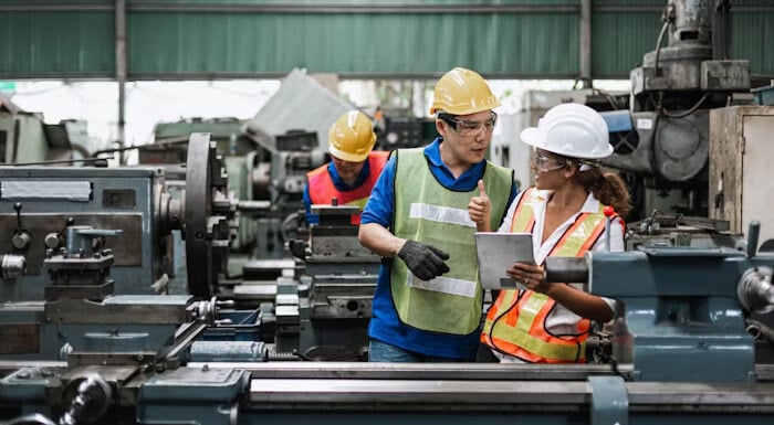 Two factory workers in safety gear discussing details on a tablet in a machine shop