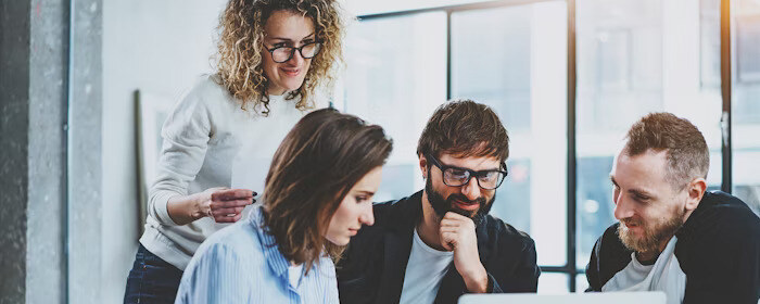 A group of coworkers gathers around a laptop, collaborating and discussing a project in a bright, modern office space