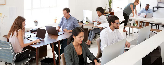 People work at desks with laptops and desktop computers in an open-plan office