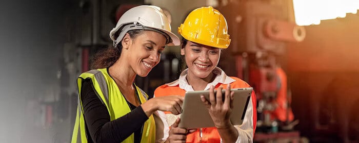 Two female workers in a factory setting wearing safety helmets and reflective vests, smiling as they look at a tablet screen, discussing work-related information