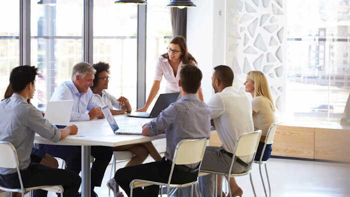 A group of colleagues sits around a table in a modern office, discussing while one woman stands and speaks to the group