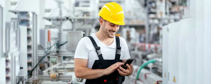 Industrial worker wearing a yellow hard hat and overalls, smiling while using a smartphone in a manufacturing facility