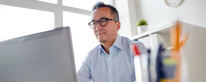 A man wearing glasses works at a desk in front of a laptop, focused on his screen in a bright office environment