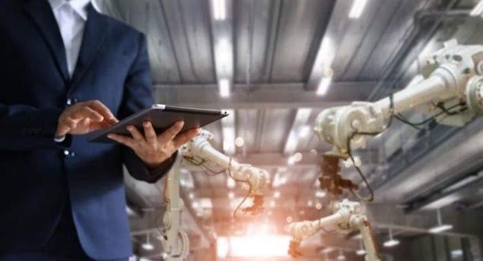 A person in a suit using a tablet in a modern factory with robotic arms in the background