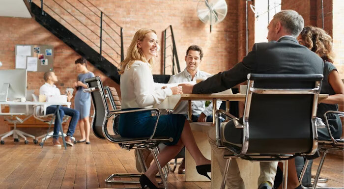 A group of people is seated around a table having a discussion in a modern office space, while others work in the background near desks and a staircase