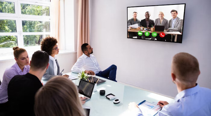 A group of people in a conference room participate in a video call with colleagues displayed on a large screen