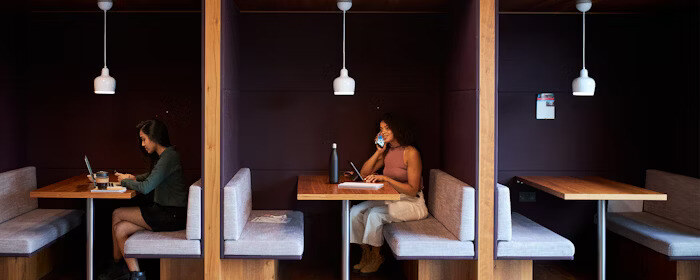 Two people work separately in booth-style seating, one using a laptop and the other talking on the phone while writing in a notebook