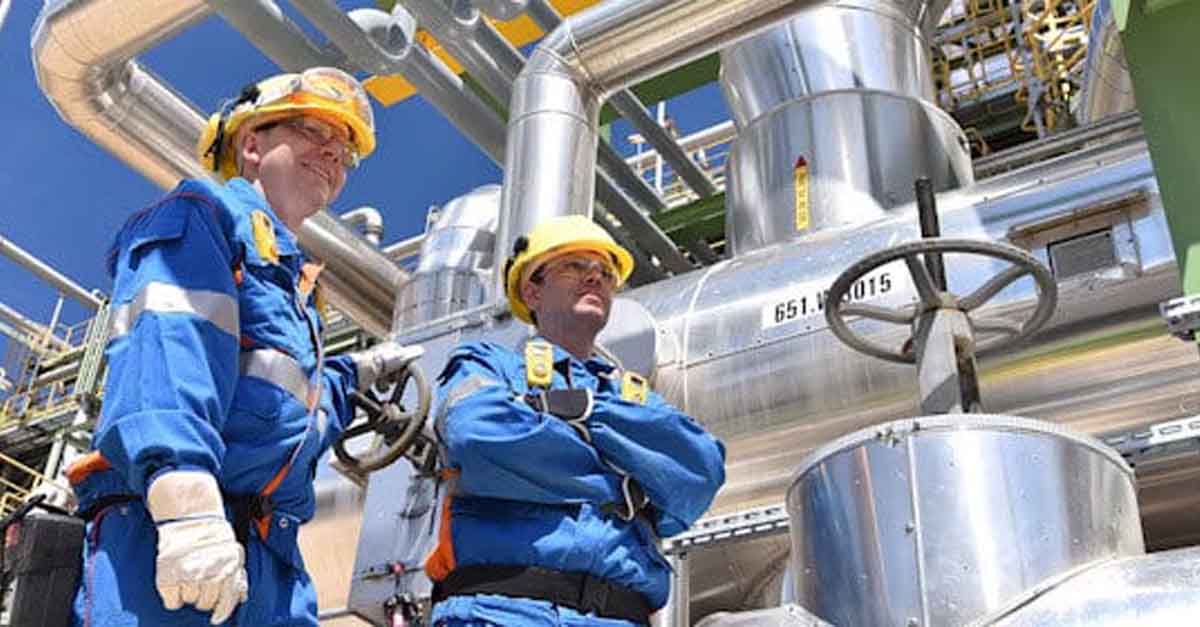 Two industrial workers standing in a refinery with pipelines and machinery