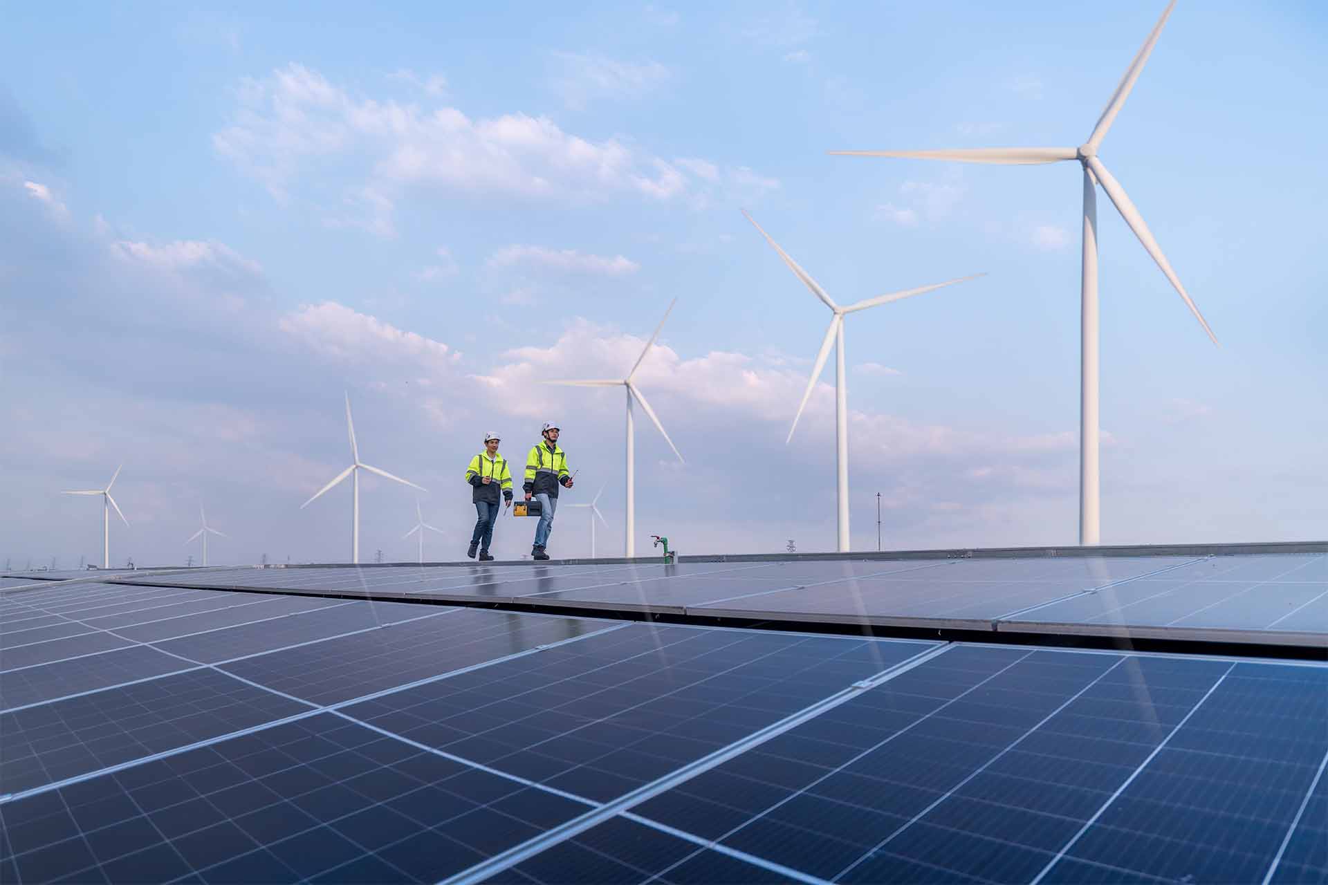 Two workers in safety gear walking on a solar farm with wind turbines in the background