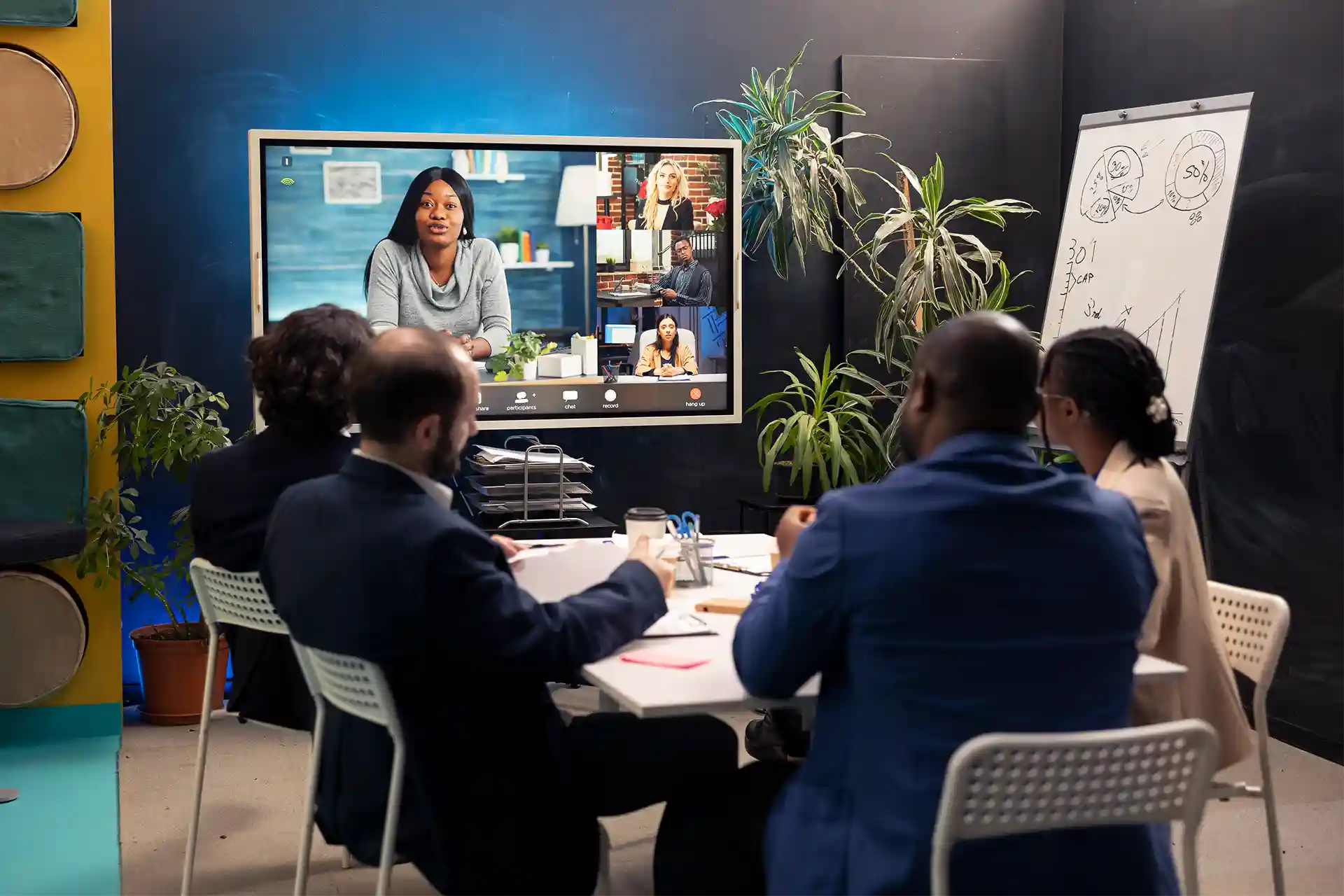 A group of professionals sits around a table in a meeting room, participating in a video conference displayed on a large screen