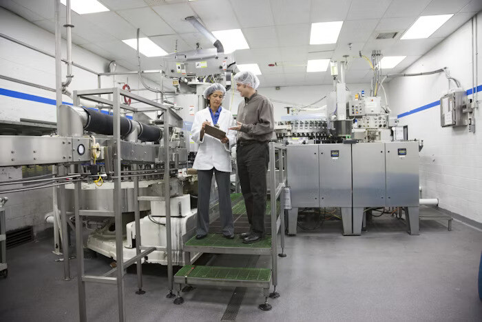 Two professionals in a manufacturing facility, wearing lab coats and hairnets, discussing and reviewing information on a clipboard near industrial machinery.