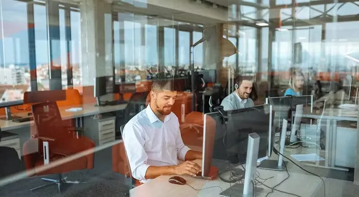 People work at computers in a modern office, with large windows providing a view of the city outside