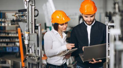 Two engineers in orange hard hats working on a laptop and tablet in a manufacturing facility