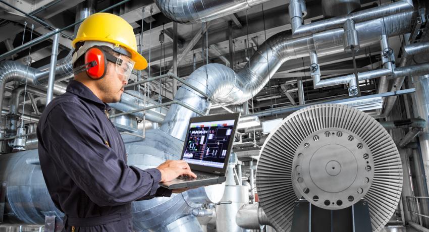 An industrial engineer in a yellow hard hat and ear protection using a laptop to monitor machinery in a factory