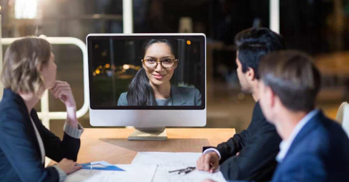 Three people in a meeting are sitting around a table, watching a woman on a computer screen during a video conference