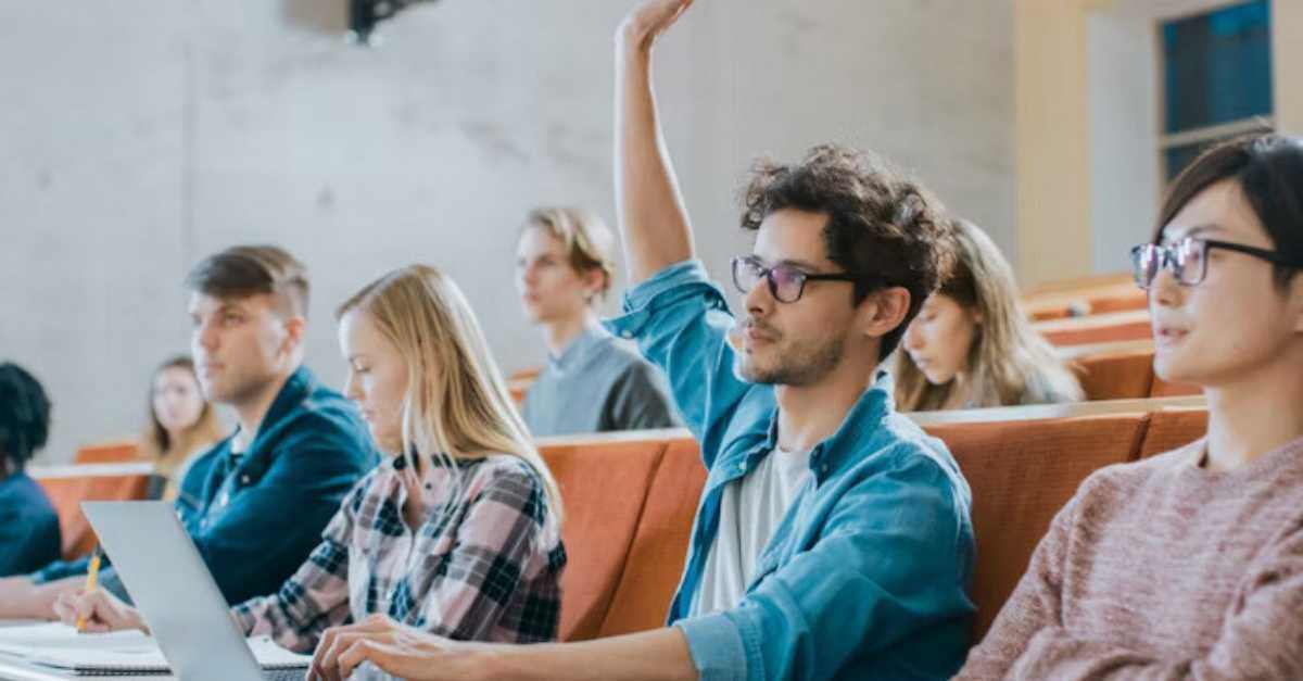 A student sitting in a lecture hall raises his hand while others around him take notes and use laptops