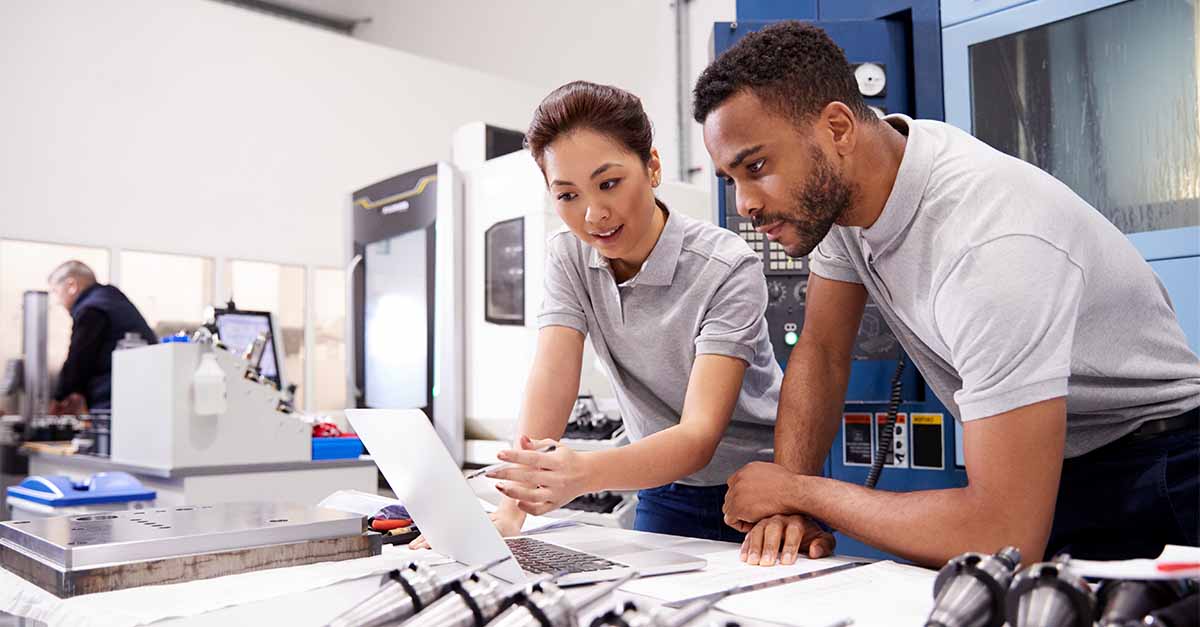 Two engineers in a manufacturing facility reviewing data on a laptop, surrounded by machinery and technical equipment.