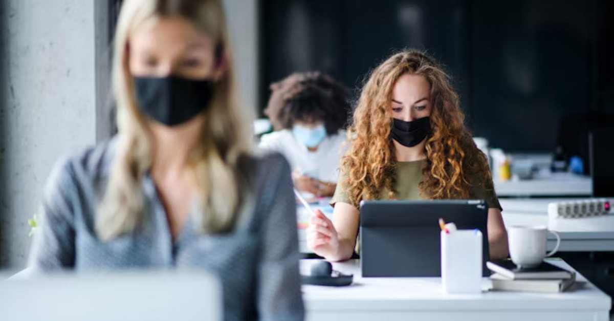 People wearing face masks work at desks in an office setting, using laptops and tablets while practicing social distancing