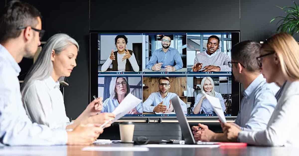 A group of people in a meeting room reviews documents while participating in a video conference with colleagues displayed on a large screen