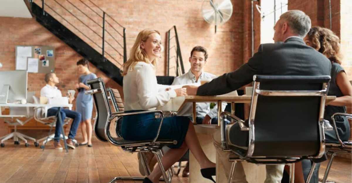 A group of people is seated around a table having a discussion in a modern office space, while others work in the background near desks and a staircase