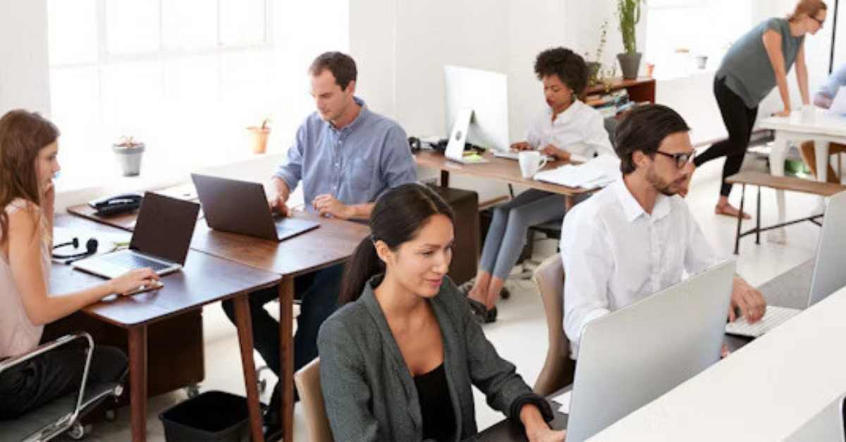 People work at desks with laptops and desktop computers in an open-plan office