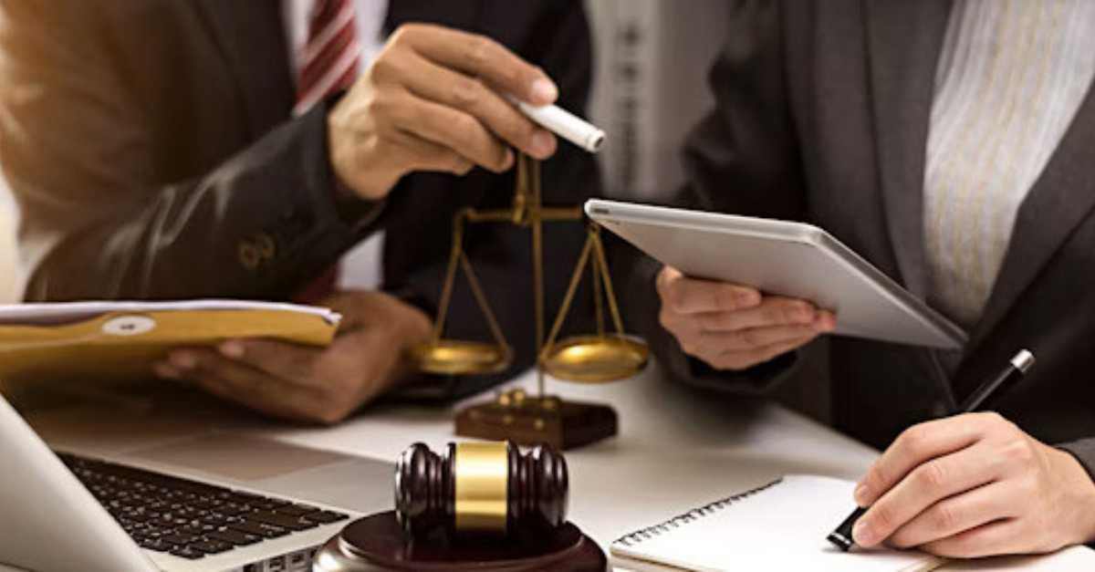 Two professionals review documents and use a tablet at a desk with legal symbols, including a gavel and scales of justice