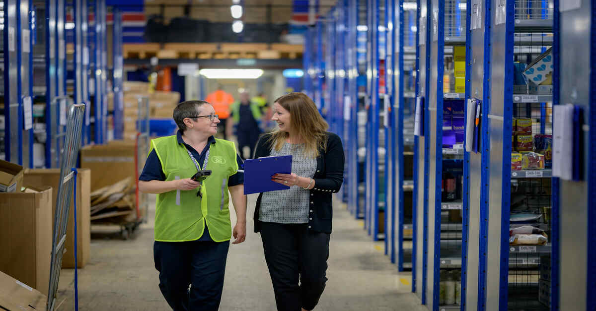 Two colleagues walking and discussing inventory in a brightly lit warehouse aisle with shelves stocked with various items.