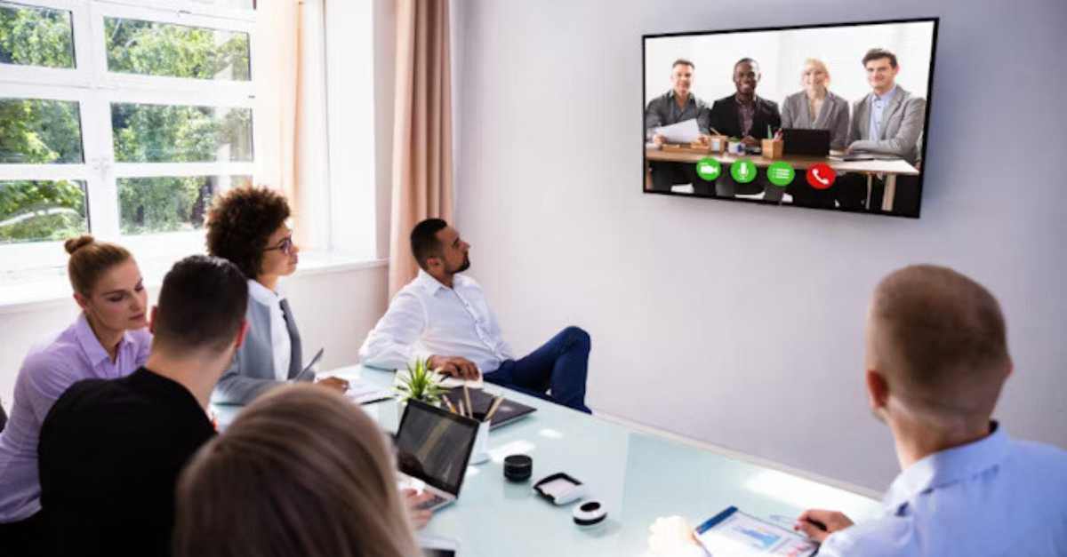 A group of people in a conference room participate in a video call with colleagues displayed on a large screen