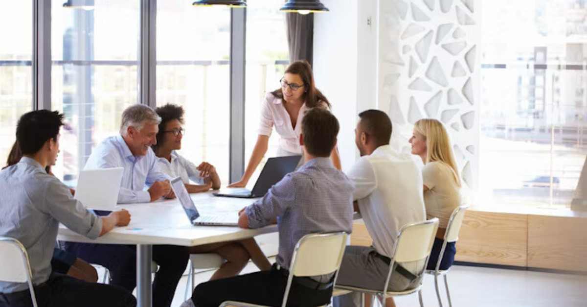 A group of colleagues sits around a table in a modern office, discussing while one woman stands and speaks to the group