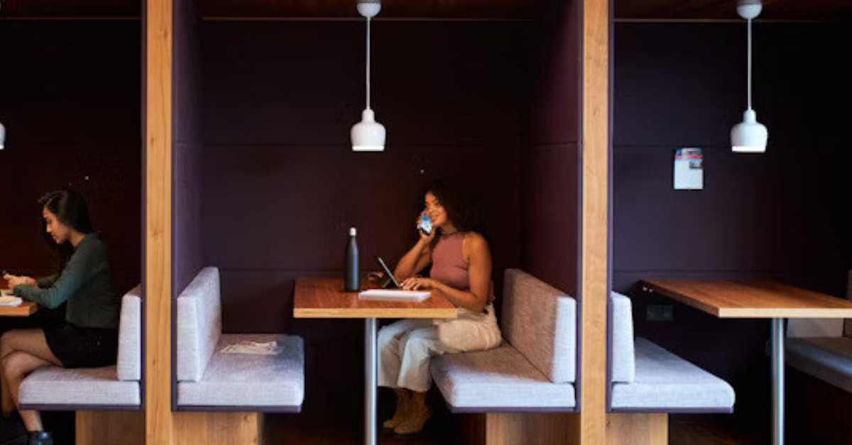 Two people work separately in booth-style seating, one using a laptop and the other talking on the phone while writing in a notebook