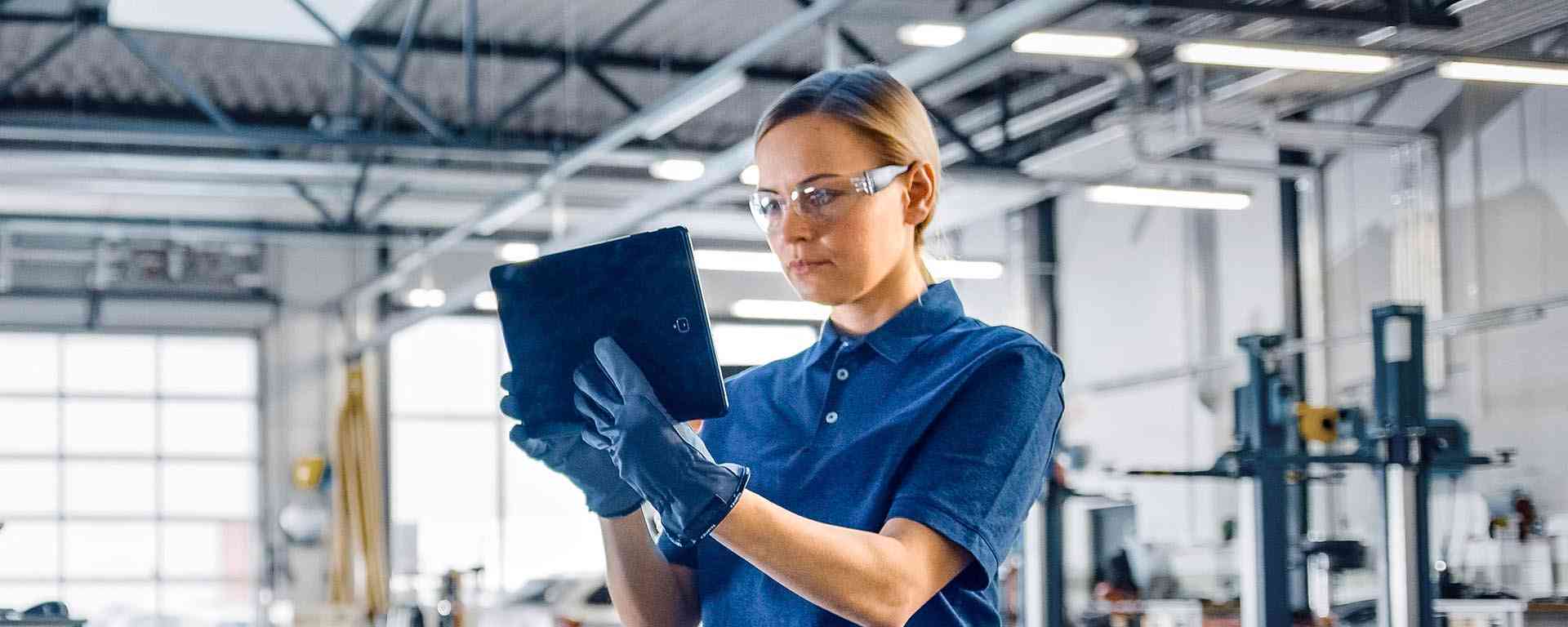 A woman wearing safety goggles and gloves, holding a tablet in a well-lit industrial or manufacturing environment